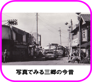 左側に、昭和41年(1966年)に設置された、斜めストライプの背面版のついた市内初の信号機と、その下に立つ制服の警官、中央に、商店の間の通りを奥から来た数台の車が、交差点の赤信号で停まっている様子が映る白黒写真（写真で見る三郷の今昔のページへリンク）