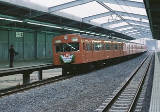 駅員が立っている手前のホームにオレンジ色の車体の電車が停車している写真