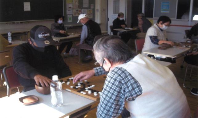 男性2名が向かい合って座り、白いベストを着た男性が白い碁石を指先でつまみ盤の上に置こうとしている囲碁の様子の写真