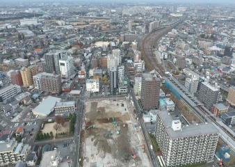 真下に見える工事現場には複数の工事車両が入り工事が進んでおり、高層ビルの間にカーブがかった線路が遠くまで続いている三郷駅周辺の様子を上空から撮った写真