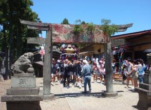 青空のもと神輿を担いだ法被姿の人たちが、神社の鳥居を通り抜けようとしている様子の写真