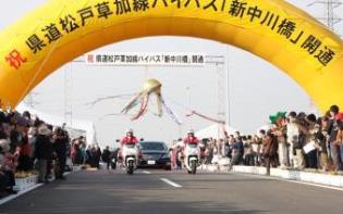 新中川橋開通イベントで、大きな黄色のアーチの下で大勢の人が見守る中、1台の黒い車と2台の白いバイクが走っている写真