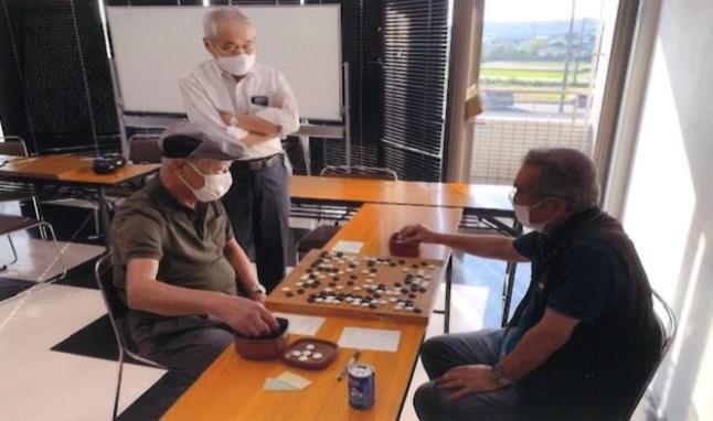 男性2名が長机の上に置かれた囲碁盤で囲碁をしており、それを白髪の男性が立って見ている写真
