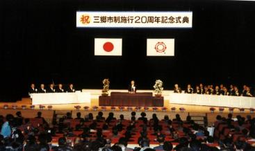 ステージに三郷市制施行20周年記念式典と書かれた横断幕とその下に、日本国旗と三郷市市章が掲げられ、中央に設置された演台で男性が挨拶をしている三郷市制施行20周年記念式典の写真