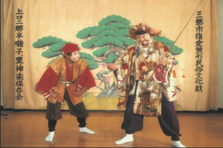 お面と衣装を身に着けて、松の絵が描かれた幕の前で里神楽を披露する2人の写真