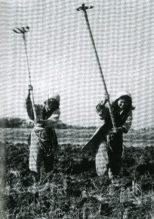 2人の女性がもんぺを着て、草が生い茂った土を万能で耕している白黒写真