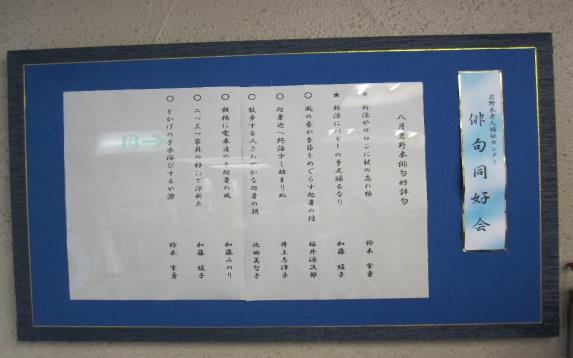 白い紙に印刷された「八月岩野木俳句好評句」8句が、黒い縁取りで背面が青の額に入れられて白い壁に掲示されている写真