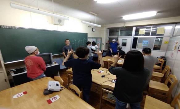 教室に木の机と椅子が置かれており、複数の参加者がその間に立って両手の中指と薬指を曲げる手話をしている手話教室の様子の写真