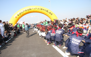 新中川橋開通イベントで、大きな黄色のアーチの下で大勢の人が見守る中、子ども達が綱引きをしている写真