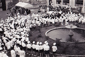 植木や竹に囲まれた噴水のある広場に多くの市民が集まり、町制施行を祝うパレードが行われている様子の白黒写真