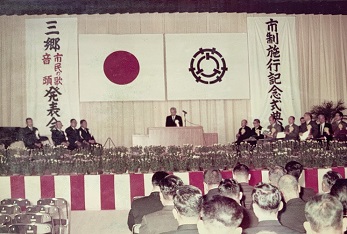 ステージ上のカーテン中央に日の丸と三郷市の市章が掲げられ、演台前で話をしている男性、両脇に関係者が座っている市制施行記念式典の様子を客席の後方からステージに向かって全体を写した写真