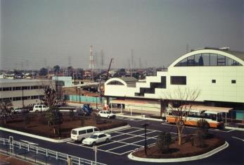 駅前のバス停にバスが停車し手前に駐車場が整備され改装した新三郷駅の駅舎の写真