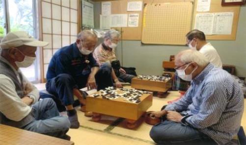 障子付きの窓と、複数の掲示物が貼られた壁のある和室に、碁盤が2台置かれ、それを挟んで高齢の男性がそれぞれ2人ずつ座り、碁盤の上に白と黒の碁石をたくさん並べて対局をしている様子と、それを手前に座った高齢男性が腕組をして見ている様子の写真