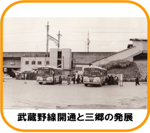 手前に停まった2台のバスのまわりにたくさんの乗客がおり、背後を通る高架鉄道の右手にはホームへ続く階段がある、開業時、昭和48年（1973年）の三郷駅前の白黒写真（武蔵野線開通と三郷の発展のページへリンク）