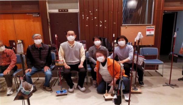 吹き矢の道具が置かれた室内の壁際付近にマスクをつけた6人の会員の方が座っており、記念撮影をしている写真