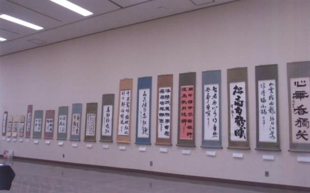 様々な書体で書かれた、たくさんの書道作品が、それぞれ色や長さの異なる掛け軸に表装され、室内の白い壁に並んで掛けられている写真