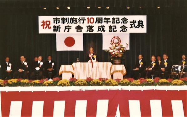 市制施行10周年を祝う式典の舞台上で演壇に立って挨拶をしている男性と、演壇の両脇に設置されたイスに座っている関係者たちの写真