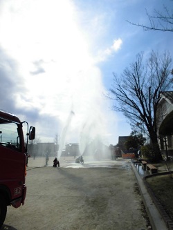左手前に消防車が停車し右側の校舎の前で放水訓練が行われている様子の写真