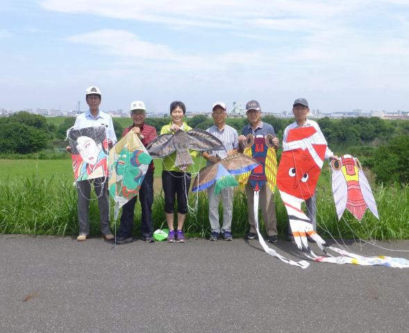 土手で会員6人が浮世絵や鳥、セミなどそれぞれが作った凧を持って立っている写真