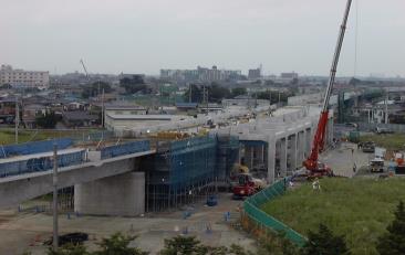 橋に工事用の網が掛けられ、右端にクレーン車が稼働している建設中のつくばエクスプレス三郷中央駅の写真