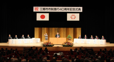 市制施行40周年記念式典で、横断幕と日本国旗、市章の旗、金屏風で飾られたステージに登壇し挨拶をしている男性と、座って話を聞く関係者の写真