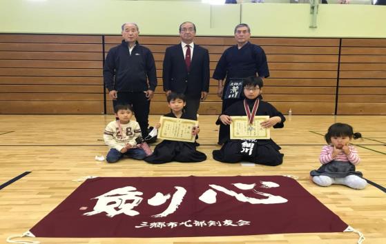 体育館で、手前の床に「剣心」と書かれた赤紫色の応援を広げ、賞状を胸の前に掲げて正面を向き、正座をしている剣道着の男の子2人と、その両脇に座る男の子と女の子、背後に立つ男性3名の集合写真