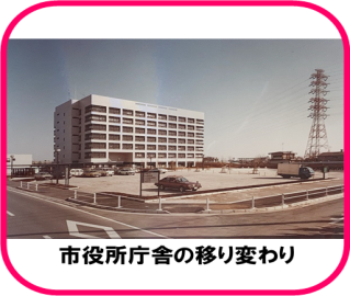 道路に面した角地に、7階建て程の白壁の建物があり、手前に車が数台停まった広い駐車場が広がって、その右奥に背の高い送電鉄塔がある、昭和58年(1983年)の三郷市役所の写真（市役所庁舎の移り変わりのページへリンク）