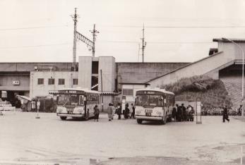 駅前の広場のバス停に2台のバスが停車し、バスに乗る為に、乗客が集まっている白黒写真