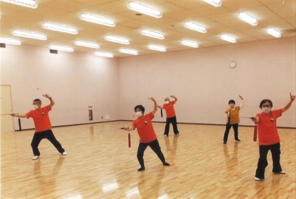 体育館の中で前後2列に並んだ、同じオレンジ色のTシャツを着た男女が、右脚を大きく前に出して立ち、剣を持った右手を前へ突き出し、左手は後ろに引いて上に挙げ、太極拳のポーズをとっている写真