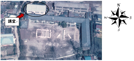 木造校舎の左側に建つ講堂を黒い丸で囲んで示している彦成小学校全体を上空から撮影した写真