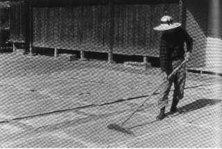 地面に莚を敷き、男性がかえし棒を使って籾を干している白黒写真