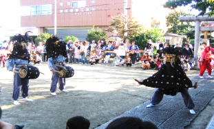 周りで多くの観客が見ている中、踊りを披露する一匹の獅子舞と、少し距離をとった所で腰につけた太鼓を叩いている2匹の獅子舞たちの写真