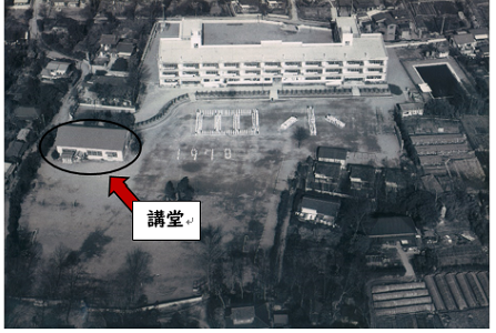 鉄筋コンクリート造りの校舎の斜め左側に建つ講堂を黒丸で示している彦成小学校全体を上空から撮影した白黒写真