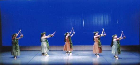 5人の女性が頭・首・手首・足首に葉っぱの飾りを付け、緑や茶色の衣装に身を包み、棒を持って踊っている写真