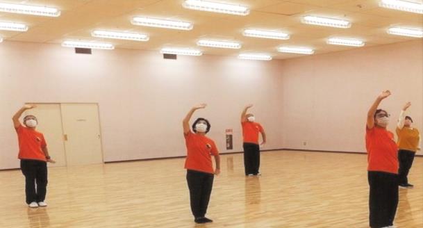 体育館の中で同じオレンジ色のTシャツを着た男女が、間隔を空けて、両足をそろえて立ち、右手が天井、左手が床を押すような気功のポーズをしている写真