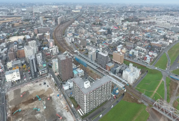 高層の建物が並び、工事現場には複数の工事車両が入り工事が進んでおり、街並みの中にカーブがかった線路が続いている三郷駅周辺の様子を上空から撮った写真