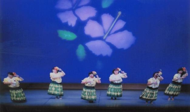 壁にハイビスカスのイラストが映された舞台上で、緑のスカートの衣装を着た6人の女性がフラダンスを披露している写真