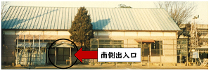 中央に1本の木が立つ左側に南側出入口を黒い丸で囲んで示している1階建ての講堂の建物外観の写真