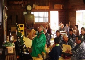神前に座りお経を転読する緑色の袈裟を着た迎攝院住職と、その後ろに座る人々の写真