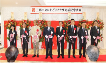 壁に紅白幕が貼られお祝いの花が並んでおり、8名の関係者が横一列に並んで立ち、テープカットを行った直後の完成記念式典の様子の写真