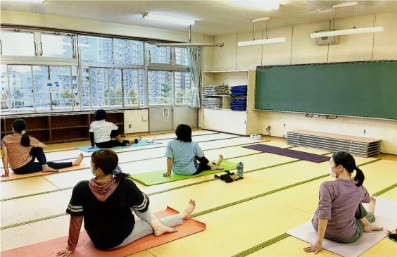 ヨガマットに右足を伸ばして座り、左足は膝を立てて右足とクロスさせ体を左側にひねるポーズをしている5名の参加者の写真
