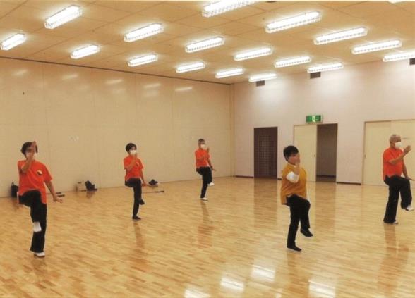 体育館の中で前後2列に並んだ、同じオレンジ色のTシャツを着た男女が、右脚の膝を腰の位置まで上げて立ち、太極拳のポーズをとっている写真