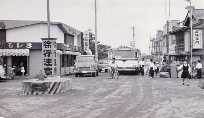 徐行注意と書かれた柱が交差点の中央に設置され、バスや車、自転車等が交差点に入ろうとしている白黒写真