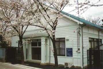 入口前に咲いている細い2本の桜と、緑色の三角屋根で白っぽい外壁の郷土資料館（現 彦成小学校講堂記念館）の写真