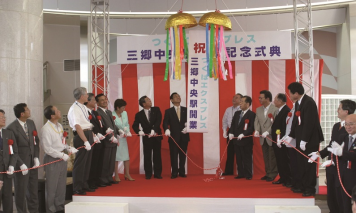 三郷中央駅開業記念式典の横断幕と紅白幕の前でたくさんの関係者が赤いテープを持ち、頭上のくす玉が割れ、三郷中央駅開業を祝う式典の様子の写真
