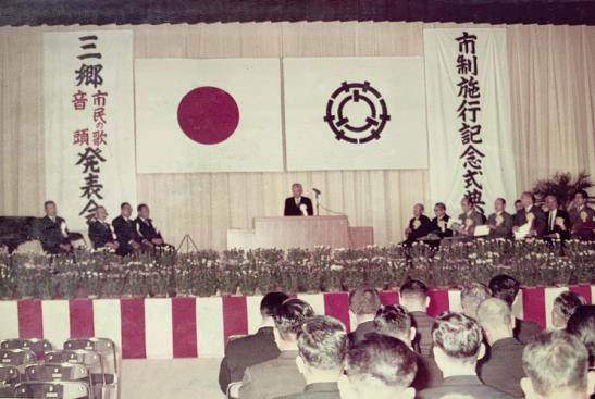 横断幕に「市制施行記念式典」と書かれ日本国旗や三郷市の市章が掲げられている舞台上で演壇に立つスーツ姿の男性と壇上に設けられた椅子に座っている関係者の方々と多くの参加者の皆さんが会場の席に着いている式典の様子の写真
