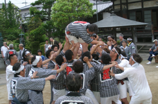 中心にいる法被姿の男性が高い位置で屈んでおり、周りにいる同じく法被姿の男性たちが中心の男性を手で支えている写真