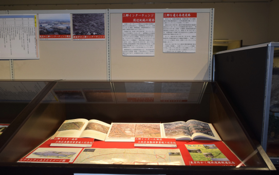 ガラス張りのケースに開かれた三郷インター土地区画整理事業竣工記念誌やパンフレットが置かれ、その後ろのボードに年表や三郷インターチェンジの写真、周辺地域の開発についての文章が展示されている写真