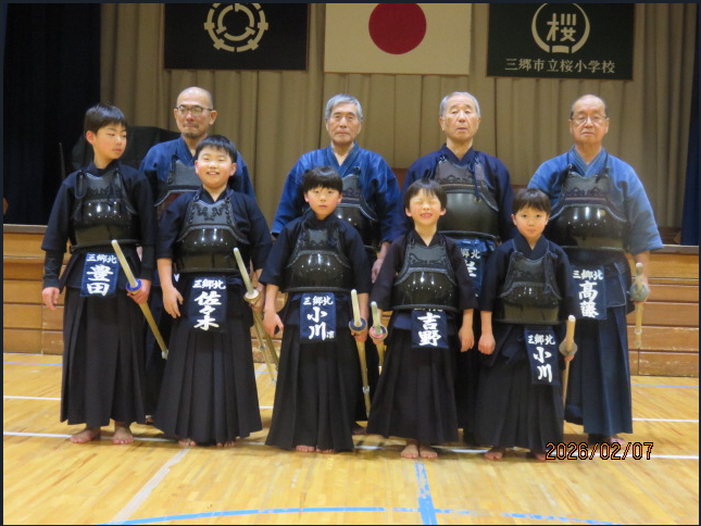 体育館の舞台の前で、剣道着に胴を身につけた子供から年配の男女が、前列は正座、後列は立って2列に並び、その手前の床に大きく「剣心」と書かれた赤紫色の応援幕が広げられている、三郷市北部剣友会全員の集合写真