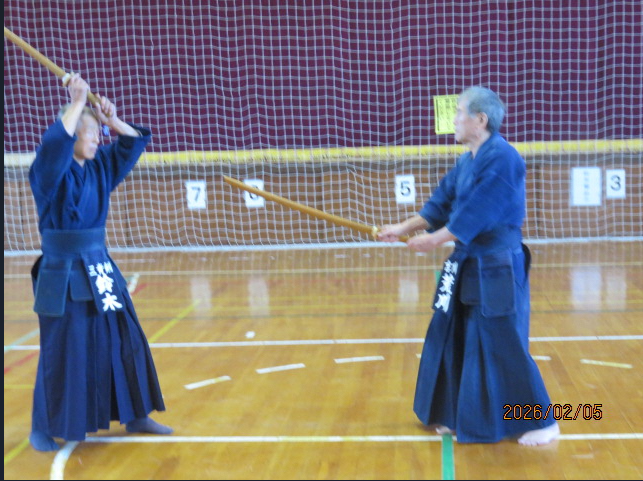 体育館の中で、舞台を背に等間隔に並んで素振りをする剣道着を着た会員たち、会員らと向かい合って竹刀を振る剣道着の男性の写真